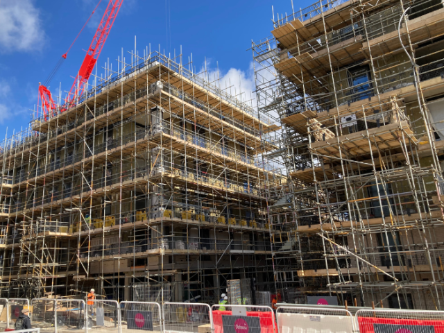 Two multi-story buildings are under construction. They are surrounded by scaffolding and the building to the left has an orange crane above it. This is set against a bight blue sky with occasional clouds. At ground level a temporary metal fence encloses the site. Two builders can be seen near the buildings in white helmets and high visibility jackets. Text reads: "Developers&rsquo; levy could support more neighbourhood improvement  schemes"