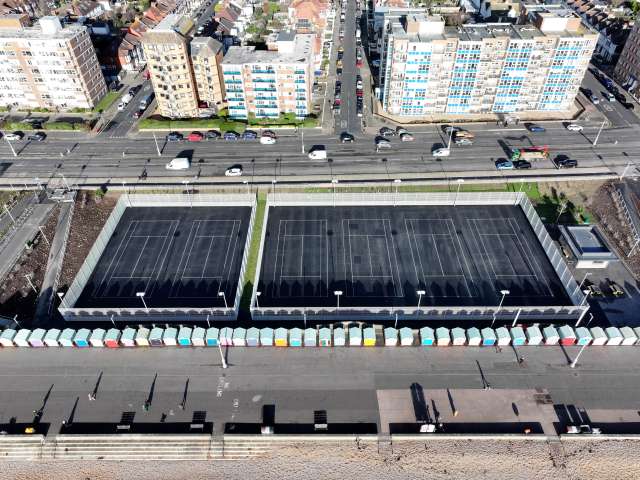 An aerial view of Hove Beach Park tennis courts shows a pair of courts to the left surrounded by white fencing,, with 4 courts to the rigth also surrounded by fencing. Beyonfd the courts is Kingswy with traffic on the road and eafront apartment blocks beyond that.