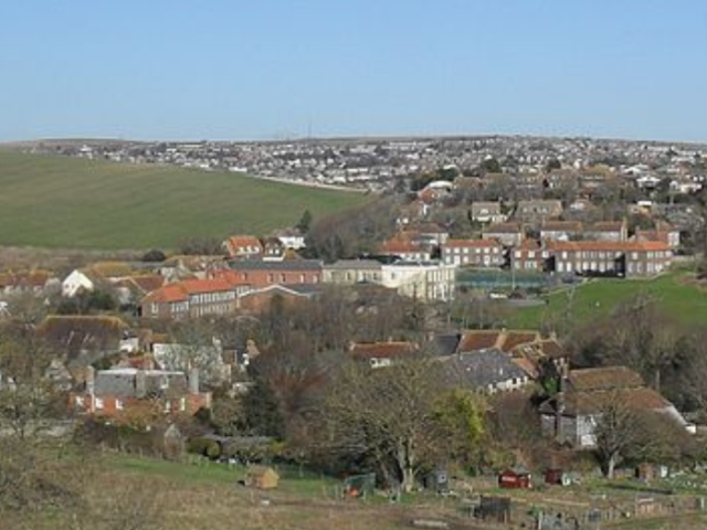 An aerial view shows Ovingdean in the foreground, with houses and other buildings showing through trees. Beyond 2 large green fields to the left and right in the background the buidlings to the east of the city stretch out.