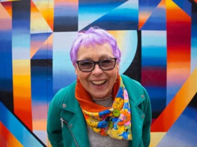A woman with purple hair and glasses smiling at the camera against a multicoloured backdrop.