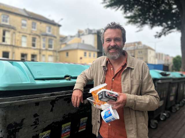 Councillor Tim Rowkins with a plastic soup pot, margarine tub, cheese tub and clear plastic tray about to be put in the communal mixed recycling bin with the light blue lid outside Hove Town Hall