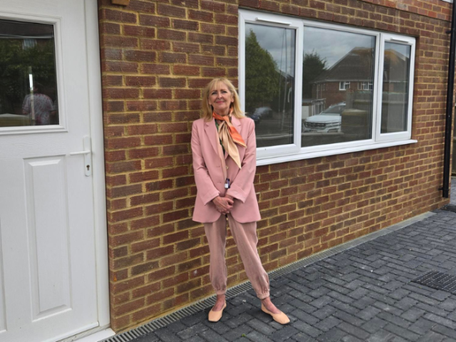 The image shows Councillor Gill Williams, clad in elegant pink ensemble, standing in front of a newly built detached house.