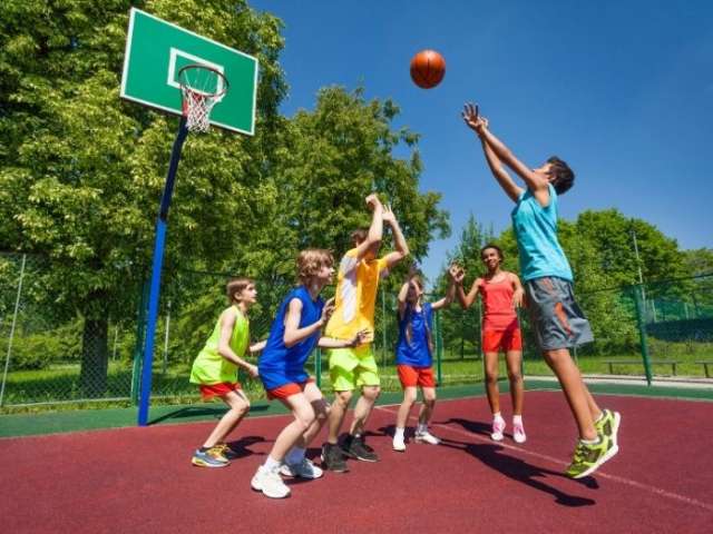 Children in multicoloured bibs playing basketball on a court outside 