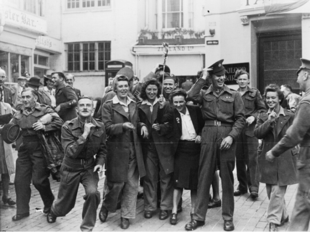 The image is a historic photo of a group consisting of women and soldiers happily celebrating VJ Day while striking a pose.