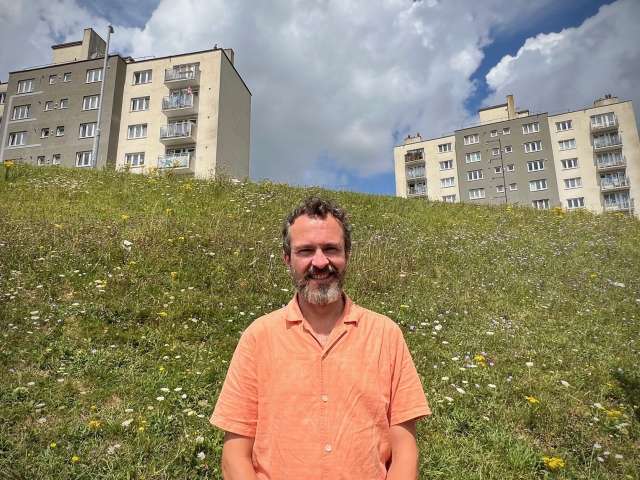 Councillor Tim Rowkins wearing an orange short sleeved shirt, with the bee bank and 2 tower blocks on the Bristol Estate behind him