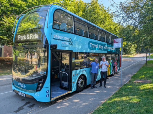 Two people standing in front of a bus with their thumbs up. The sign on the bus says 'Park and Ride'
