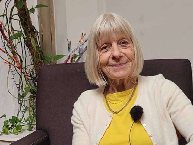  A photograph of local resident Jackie, who has shoulder length grey hair and is wearing a yellow top and white cardigan, and is sitting on a brown arm chair.