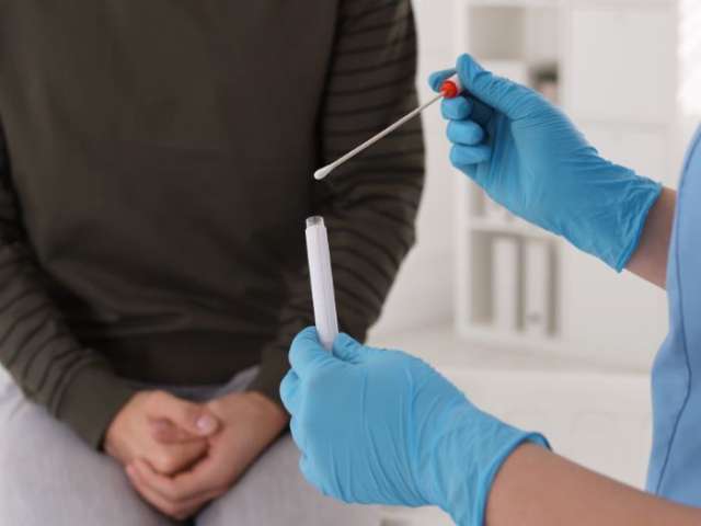 A close up shot of a healthcare professional holding a swab and in the background a person sat with their aims help in their lap.