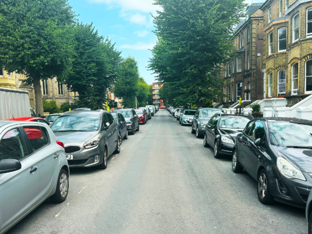 A photo of a road with parked cars on either side and trees in the background