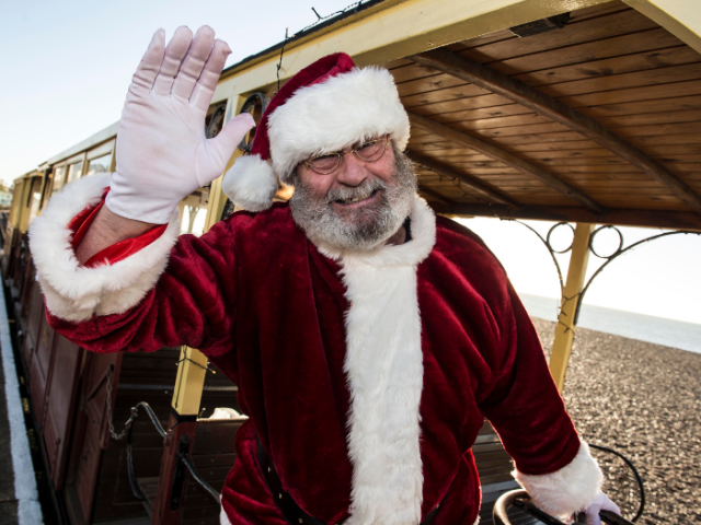 Photo of a waving man in a red and white Santa outfit with a light yellow train behind him.