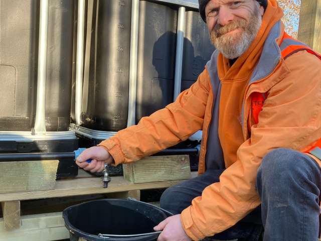 Jake wearing an orange jacket and grey woollen hat, filling up a bucket from the water saving station at Falmer allotments