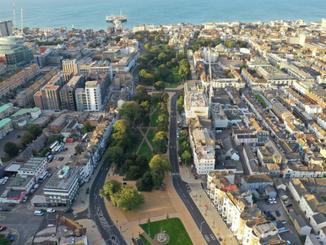 A picture from above of a city with roads. trees and buildings with the sea and a pier in the background