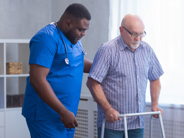 A caregiver in blue scrubs assisting an older person using a walker