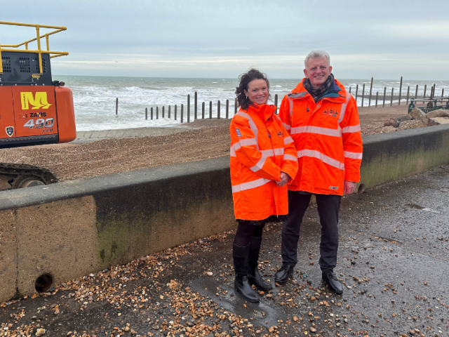 Two people in hi viz jackets standing in front of a beach, a digger and timber groynes
