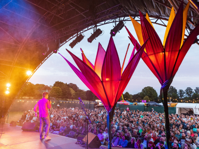 Photo looking from the back of an outdoor stage where there is a man in a pink shirt performing to a crowd of people sitting outside.