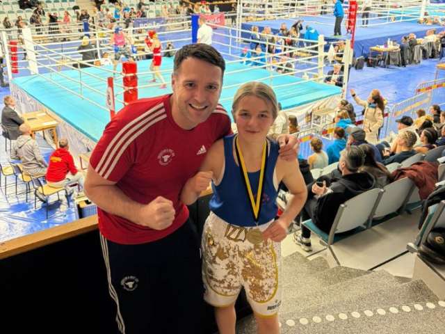 A young female boxer and her male coach stood in front of a boxing ring with their fists up 
