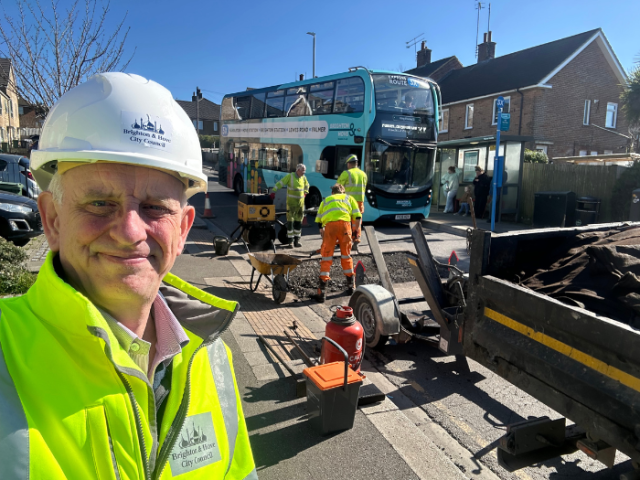 A man in a high viz jacket and hard hat posing in front of a group of men in similar clothing working on a road with a bus in the background