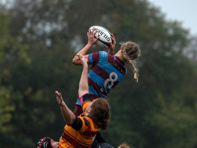 A player in maroon and blue catches a ball while being challenged by a player in orange. Neither are facing the camera and both are off the ground.