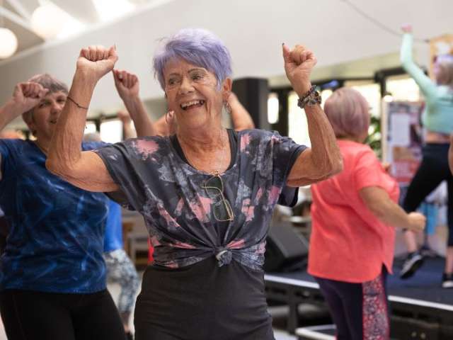 A smiling older woman with purple hair dancing in an exercise class 