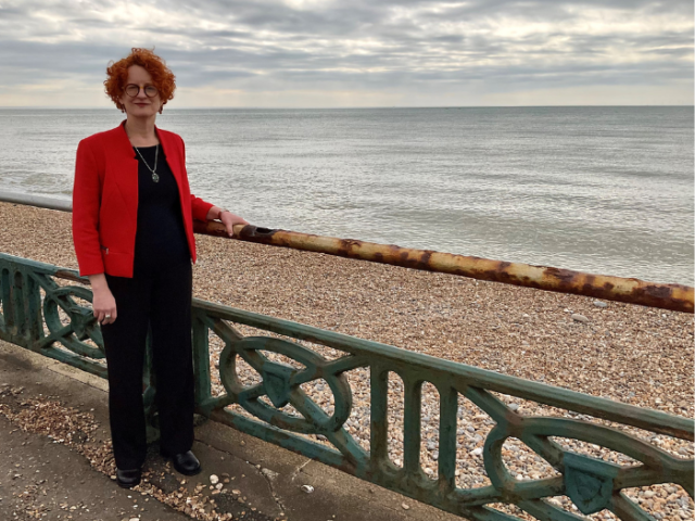 Woman with red curly hair and a red jacket stood in front of railings on the seafront.