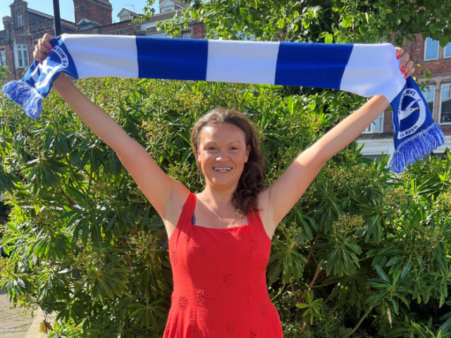 Councillor Bella Sankey, in a red summer dress, smiling and holding a blue and white Brighton & Hove Albion scarf above her head