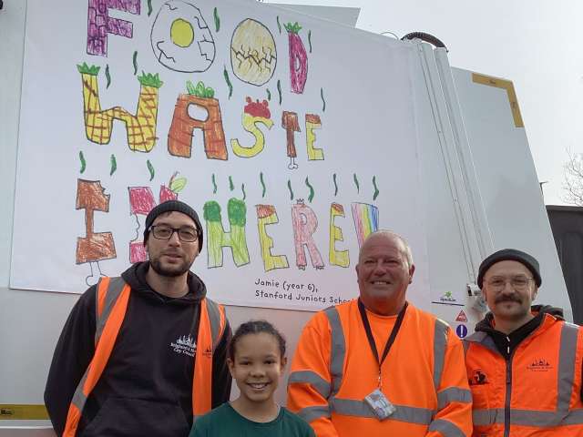 Pupil Jamie with 3 crew members standing in front of a refuse truck with her food waste design on the side. The design is made up of letters in the shape of different types of food which spell out 'Food Waste In Here.'