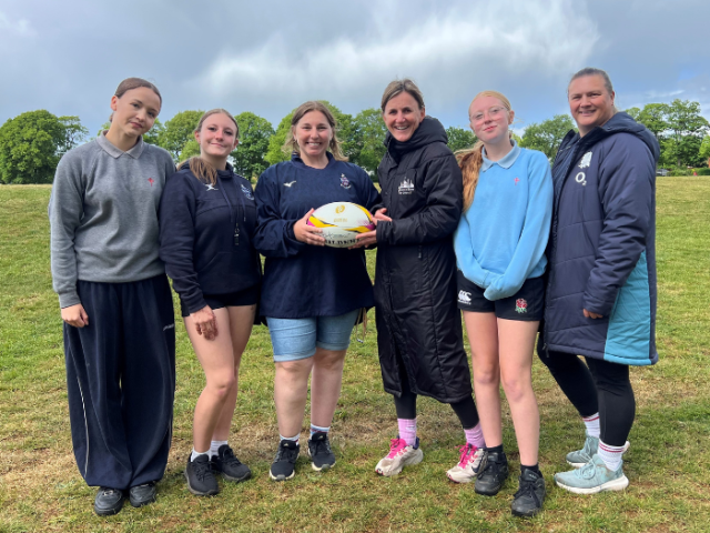 Three teenage girls and three women in a team photo pose on a rugby pitch, with the middle two holding a rugby ball between them