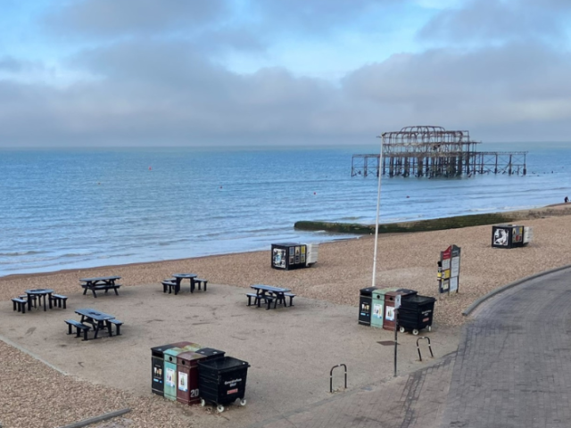 A pebbled beach in the foreground, with benches and waste bins, with the sea behind and Brighton's West Pier on the right