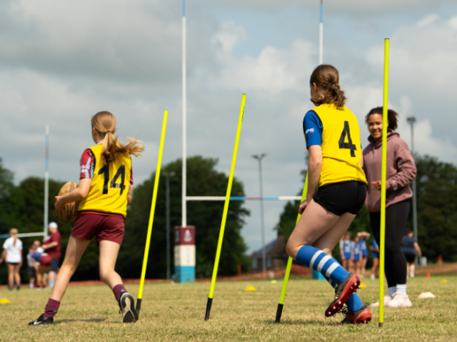 Two girls with their backs to the camera, one carrying a rugby ball and both wearing yellow bibs, weave between yellow poles while being encouraged by a women looking on