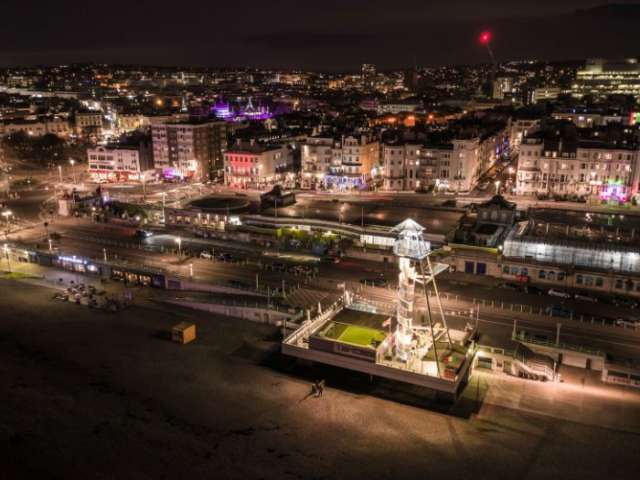 A nighttime aerial view of Brighton seafront and city centre, showing the beach, promenade, roads and brightly lit buildings stretching across the shoreline.