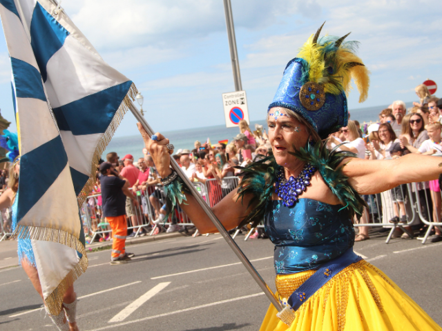 An image of a person dressed in colourful clothing holding a flag while taking part in a parade