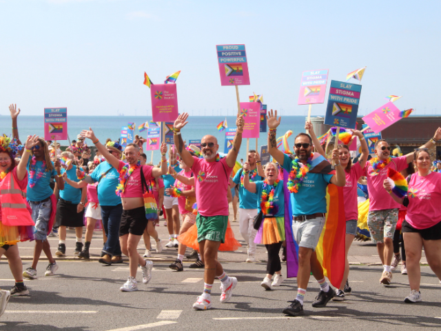 A photo of a brightly dressed group of people waving and dancing during Pride march, on Hove's seafront.