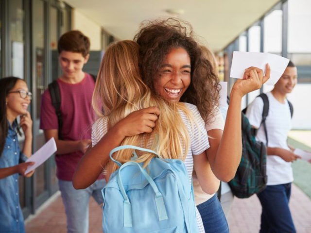 A smiling student in a school hallway hugs a friend while holding a piece of paper