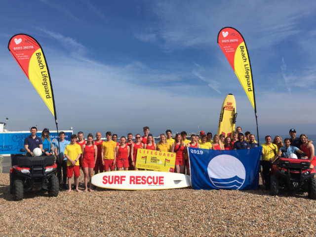 Photograph of a group of lifeguards standing on the pebbles on Brighton Beach with the sea and a blue sky backdrop. 