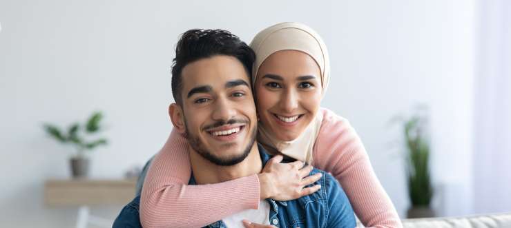2 adults smiling. One adult is hugging the other adult around the shoulders from behind