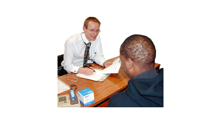 A person in a white shirt and tie with a stethoscope sitting at a table with a book and a pen. In front of the person is a person in a dark hoodie.