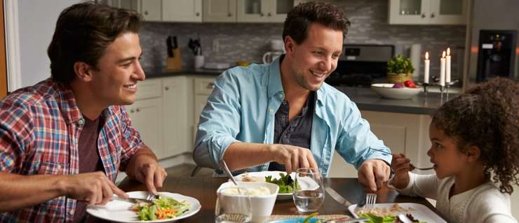 family of 2 adult men and one child eating food together at a table in a kitchen