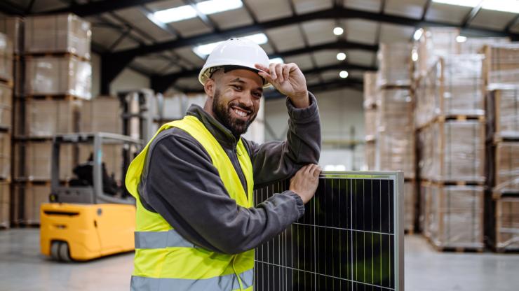 A person is standing in a warehouse wearing a yellow high vis jacket and a hard hat, they are smiling at the camera while tipping their hat.