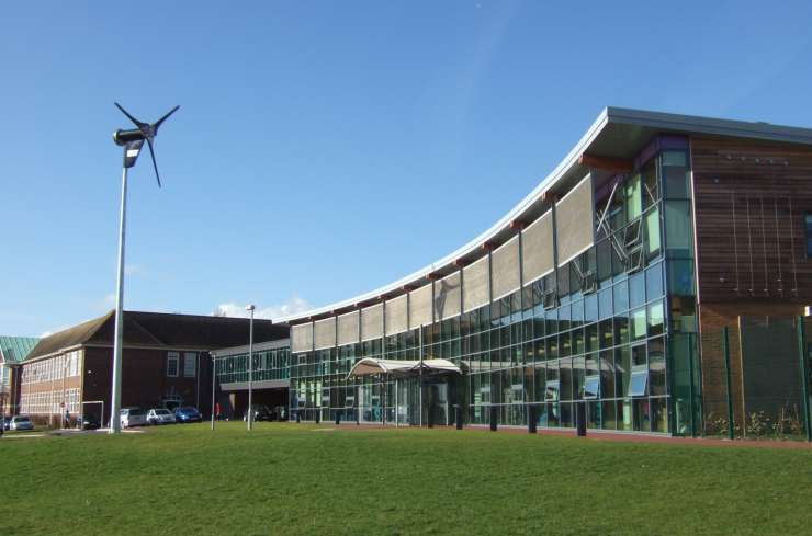 Photograph of a school building from a distance, with a green field in the foreground and a blue sky. 