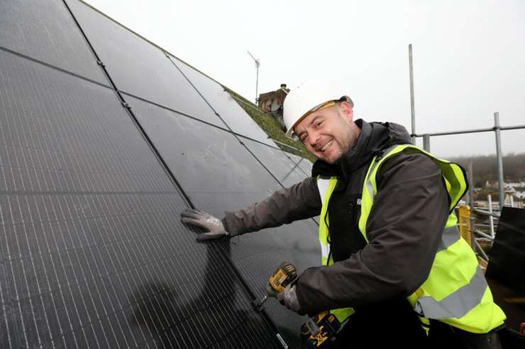 A person installing solar pannels on a roof, they are wearing a hard hat and a high vis vest.