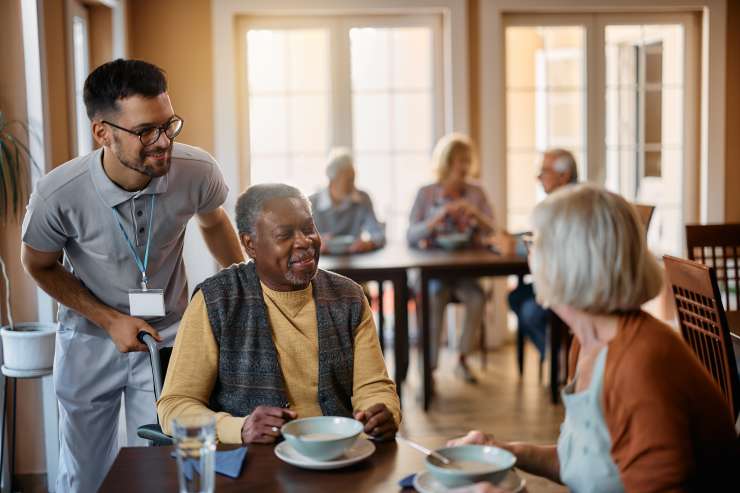 A pair of people sitting at a table eating soup, a carer is standing behind them