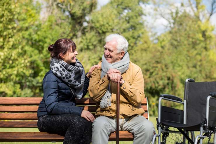 Two people sitting on a bench outside, one has a walking stick and a wheelchair