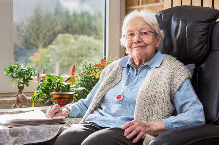 An older person sat in a highbacked chair with a alarm pendent around their neck