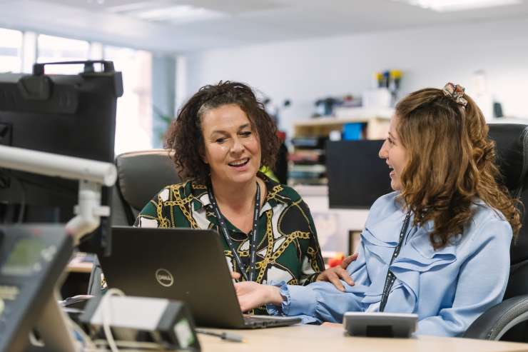 2 people sat at a desk with an open laptop, happily communicating
