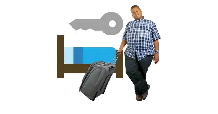 Person holding suitcase next to a bed and key to represent them staying in a hotel or somewhere temporarily.