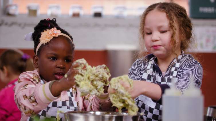 2 young children sitting at a table with their hands in a bowl of food